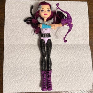 May include: A doll with purple hair, wearing a light purple top, black leggings, and purple boots. The doll has a silver collar and a blue arm band. It is holding a purple bow and arrow.