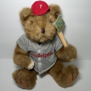 May include: A brown teddy bear wearing a grey baseball jersey with "Philadelphia" embroidered in red. The bear is wearing a red baseball cap with a white "P" and holding a wooden baseball bat. A tag reads "American Baseball Favorite Pastime".