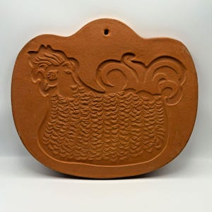 Può includere: Uno stampo in ceramica color terracotta con un design in rilievo raffigurante un gallo. Lo stampo ha una forma arrotondata simile a una zucca con un piccolo foro nella parte superiore per appenderlo. Il gallo è raffigurato di profilo.