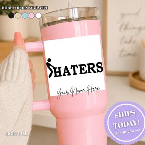 May include: A light pink tumbler with a clear lid and a handle. The front features a white rectangular label with a black graphic of a stick figure and the text HATERS. Below the graphic, the text Your Name Here is printed in black script.