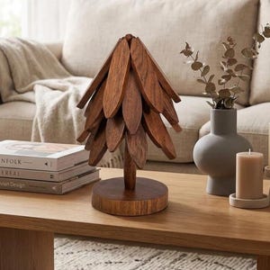 May include: A decorative wooden sculpture shaped like a pine tree with layered, pointed brown leaves on a circular base, positioned next to a stack of three books titled Kinfolk, Cereal, and Scandinavian Design, a gray vase, and two pillar candles.