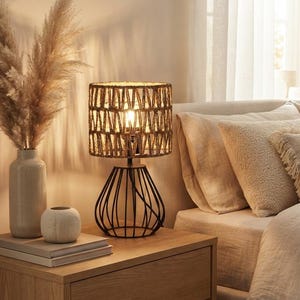 May include: A table lamp with a cylindrical shade wrapped in natural woven fibers and a black metal wire base. The lamp sits on a wooden nightstand next to a tall ceramic vase containing dried pampas grass and a small, round, textured white object.