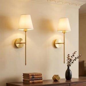 May include: Two wall-mounted lamps featuring a brushed gold finish, a slender vertical rod, and a tapered off-white fabric shade. The fixtures are mounted on circular gold backplates and emit a warm glow against the wall surface.