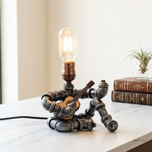 May include: A table lamp constructed from dark gray metallic plumbing pipes, shaped like a figure sitting down and playing a small wooden acoustic guitar. The lamp features a copper-colored socket holding a clear, elongated Edison-style light bulb.