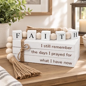 May include: A stack of three white rectangular blocks with brown text reading I still remember the days I prayed for what I have now. On top are five white cubes spelling FAITH, accented by a natural jute rope, a tassel, and wooden beads.