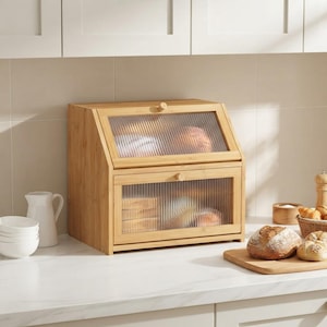 May include: A two-tier bamboo bread box with a natural light wood finish. The unit features two drop-down doors with fluted, semi-transparent glass panels and small round wooden knobs, allowing for a partial view of the bread stored inside.