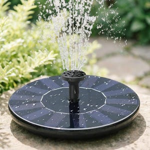 Op de afbeelding: Een fontein op zonne-energie met opspuitend water. De fontein heeft een zwarte, cirkelvormige basis met zonnepanelen en een centrale sproeier. De achtergrond toont groen gebladerte en een stenen oppervlak.