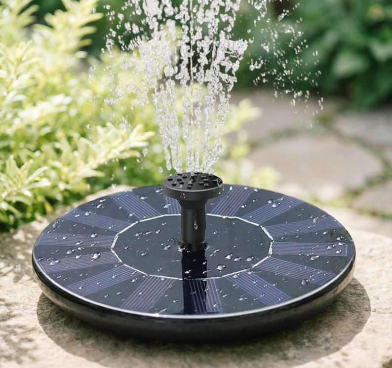 Op de afbeelding: Een fontein op zonne-energie met opspuitend water. De fontein heeft een zwarte, cirkelvormige basis met zonnepanelen en een centrale sproeier. De achtergrond toont groen gebladerte en een stenen oppervlak.