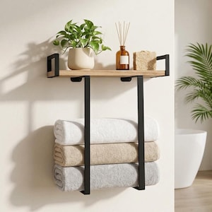 May include: A wall-mounted storage unit featuring a light wood shelf supported by black metal brackets. The shelf holds a potted green plant, a glass reed diffuser, and a textured block. Below, three folded towels in white, beige, and gray are stacked.