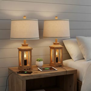 May include: Two wooden bedside lamps with beige shades and black trim. The lamps have a lantern-style base with a visible lightbulb. A small potted plant and charging devices are on the wooden nightstand. The lamps are on a wooden nightstand.