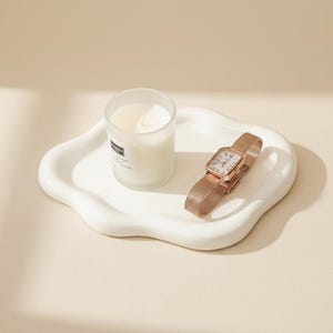 May include: A white, cloud-shaped ceramic tray holds a lit candle in a clear glass and a rose gold wristwatch with a square face and mesh band. The candle has a white label. The watch is approximately 1 inch wide.