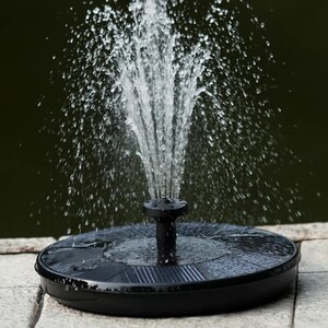 Op de afbeelding: Een zwarte waterfontein op zonne-energie met opspattend water. De fontein heeft een ronde basis met een zonnepaneel en een centrale sproeier. Het water spuit in een fonteinvorm. De achtergrond is donkergroen.