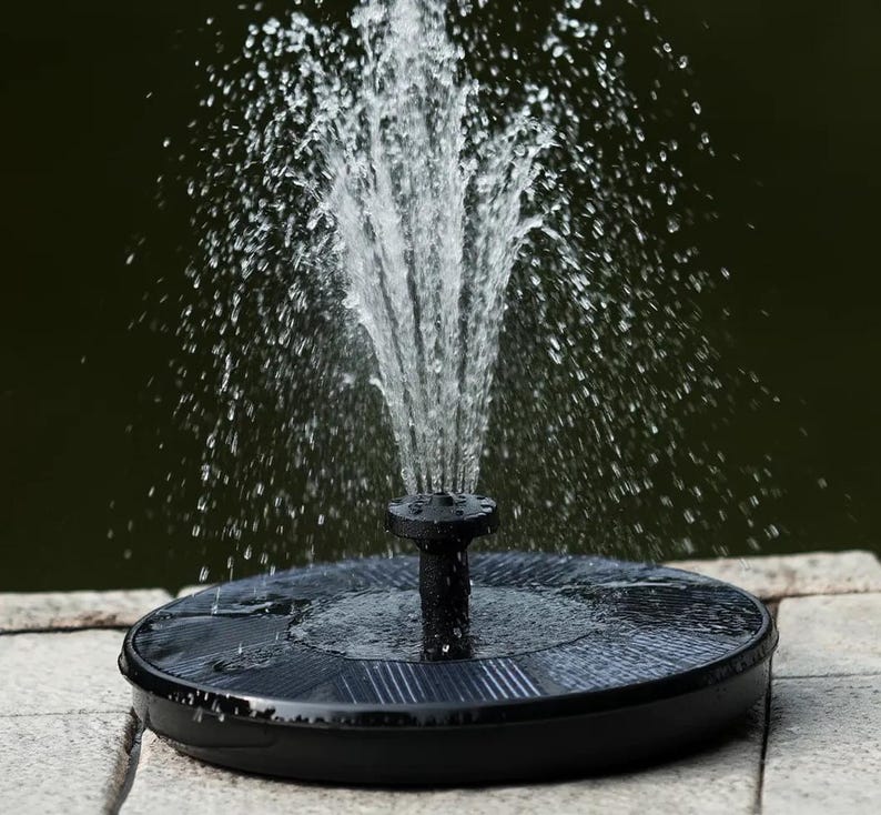 Op de afbeelding: Een zwarte waterfontein op zonne-energie met opspattend water. De fontein heeft een ronde basis met een zonnepaneel en een centrale sproeier. Het water spuit in een fonteinvorm. De achtergrond is donkergroen.