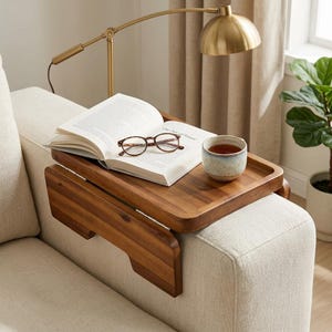 May include: A rectangular wooden sofa arm tray with hinged side panels that drape over the armrest. The tray surface holds an open book with visible text, a pair of brown-rimmed reading glasses, and a speckled ceramic mug containing dark liquid.