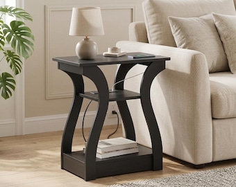 May include: A black end table with a rectangular top, a middle shelf, and a bottom shelf. The table features curved side supports and a built-in power outlet on the middle shelf. A beige lamp sits on the top surface, and books are on the bottom shelf.