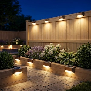 May include: Outdoor scene at dusk featuring a wooden fence and flower beds illuminated by solar-powered lights. The lights are mounted on the fence and the wooden planters, casting a warm glow on the plants and stone patio.