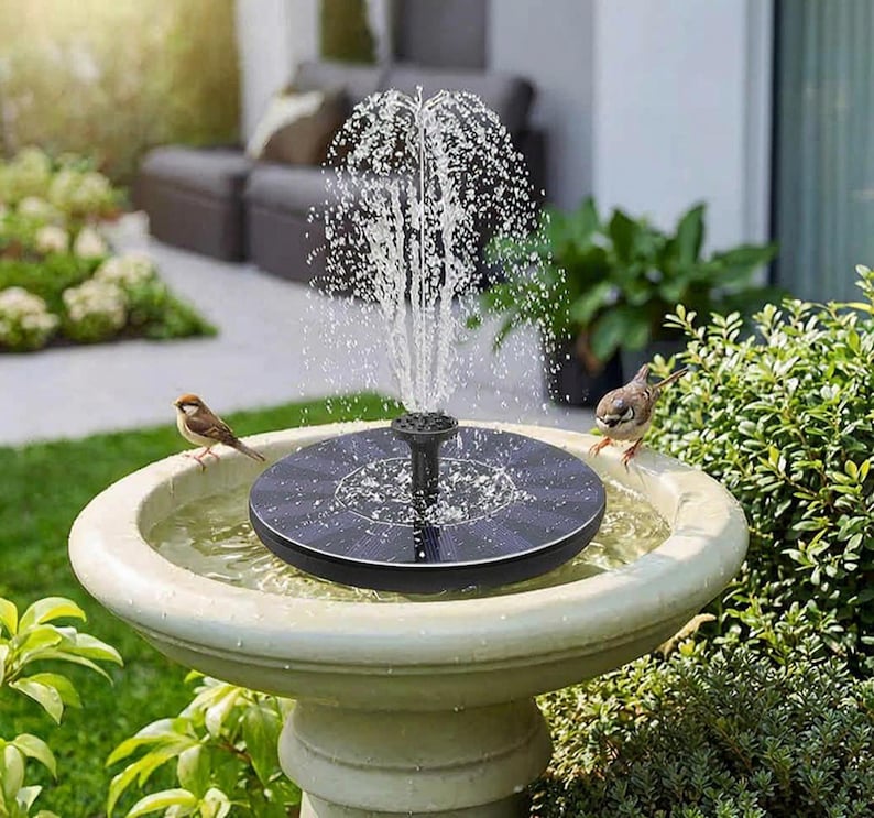 Op de afbeelding: Een fontein op zonne-energie in een stenen vogelbad. De fontein spuit water omhoog en cre&euml;ert een kleine waterpartij. Twee kleine vogels zitten op de rand van het vogelbad, dicht bij het water.