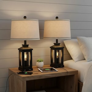 May include: Two black bedside lamps with beige fabric shades and a lantern-style base. Each lamp has a charging port and is placed on a wooden nightstand. A small succulent in a white pot and a tablet are also on the nightstand.