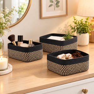 May include: Three rectangular storage baskets made of woven rope with a natural tan and black color pattern. Each basket features a solid black top rim and a base constructed from alternating tan and black rope coils, providing a structured design.