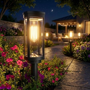 May include: Black outdoor pathway lights with a clear glass enclosure and a vintage-style Edison bulb. The lights are set in a garden with pink and purple flowers, a stone pathway, and a patio area in the background. The scene is lit at night.