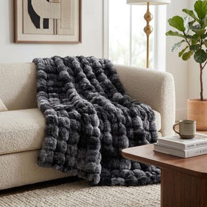 May include: A plush throw blanket featuring a textured, three-dimensional pom-pom or bubble pattern in a gradient of charcoal gray, slate, and black tones, draped over a cream-colored sofa with a soft, nubby fabric finish.