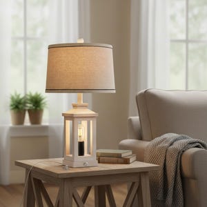 May include: A white lantern-style table lamp with a beige lampshade and a black trim. The lamp sits on a wooden side table with a light bulb visible inside. The lamp is plugged in and is next to a stack of books.