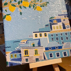 May include: A square canvas painting of an Italian coastal town. The artwork shows blue buildings with white and yellow details, set against a light blue sky with lemon tree branches and yellow lemons. The painting is displayed on a small wooden easel.