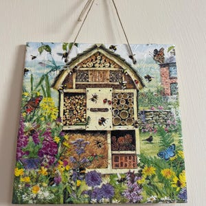 May include: A square, decorative wooden plaque featuring a detailed illustration of a bug hotel. The artwork includes a variety of insects, flowers, and a small house. The plaque is suspended by a natural twine cord.