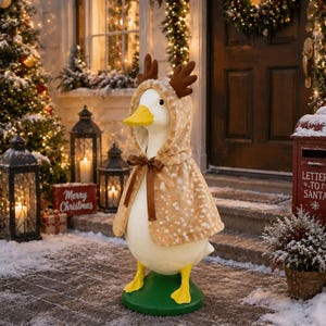 May include: A white goose figurine standing on a green base, wearing a tan hooded cape with a white spotted pattern and brown plush reindeer antlers attached to the hood. The cape is secured at the neck with a thin brown satin ribbon bow.