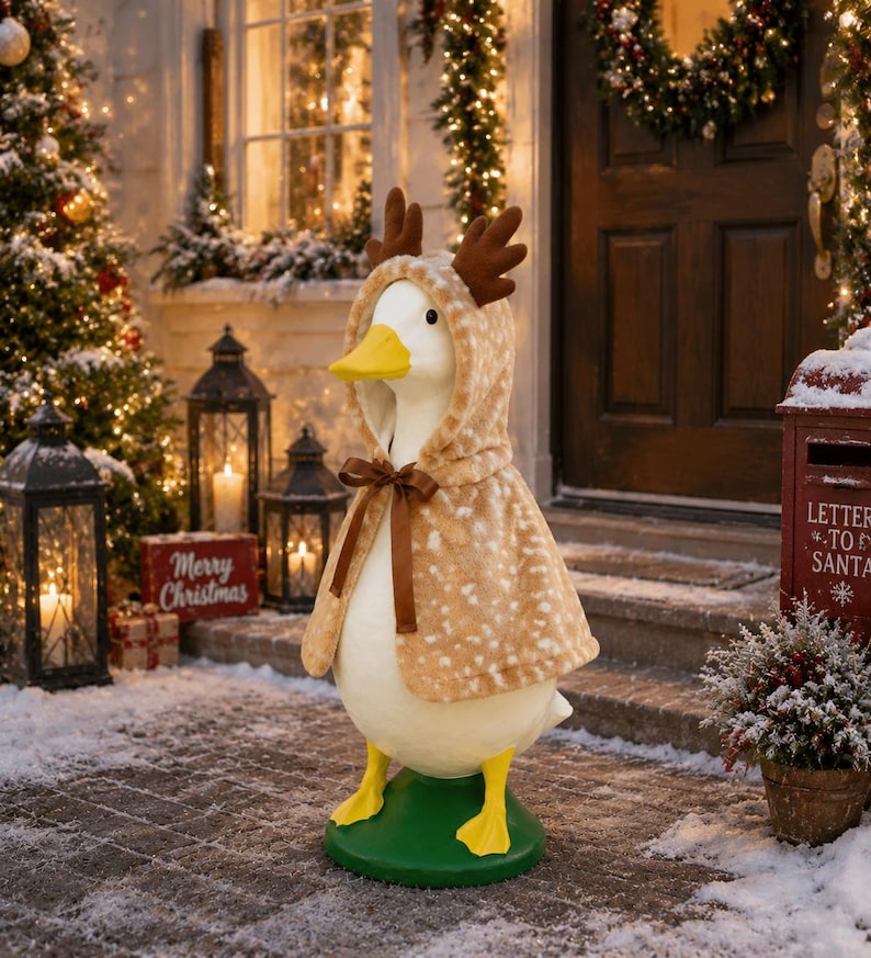 May include: A white goose figurine standing on a green base, wearing a tan hooded cape with a white spotted pattern and brown plush reindeer antlers attached to the hood. The cape is secured at the neck with a thin brown satin ribbon bow.