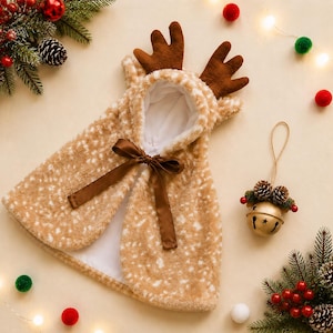 May include: A tan and white spotted faux fur hooded cape featuring brown plush reindeer antlers on the hood and a brown satin ribbon tie closure at the neck. The interior is lined with white fabric, and the exterior has a soft, textured finish.