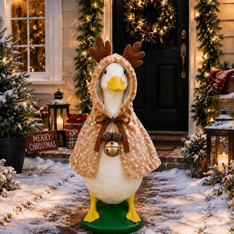 May include: A white goose figurine wearing a tan, spotted hooded cape with brown plush reindeer antlers. The cape is secured with a brown ribbon bow featuring a gold jingle bell and two small pinecones. The goose stands on a green base.