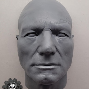 May include: A gray, sculpted bust of a human face, featuring detailed wrinkles and facial features. The sculpture has a bald head and a stern expression. A logo with a skull and beard is in the lower left corner.