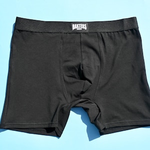Op de afbeelding: Zwarte boxershort met een zwarte tailleband. De tailleband heeft de witte tekst "BAKTERS". Het ondergoed is gemaakt van een zacht materiaal en wordt gepresenteerd op een lichtblauwe achtergrond.