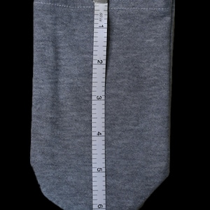 May include: A grey fabric pouch with a rounded bottom, approximately 18 cm in length, is shown against a black background. A measuring tape is placed next to the pouch, indicating its size. The pouch appears to be made of a soft, knit material.