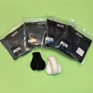 Puede incluir: Cuatro bolsas de plástico transparente selladas que contienen boxers oscuros, cada una etiquetada con el logotipo de Baxters Boxers. Frente a las bolsas hay dos bolsas de tela suave y redondeada, una negra y otra blanca, con forma de testículos.