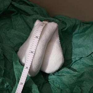 May include: A white, folded fabric item is measured with a white measuring tape. The tape shows measurements in inches. The fabric is resting on a bed of dark green, crinkled paper. The measurement is shown in cm.