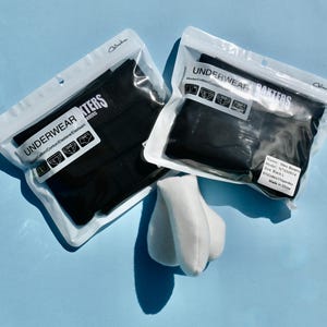 Puede incluir: Dos paquetes de ropa interior negra etiquetados "UNDERWEAR BOXERS" en envases de plástico transparente. Los paquetes están sobre una superficie azul claro. Un objeto de felpa blanca está delante de los paquetes.
