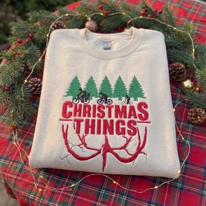 May include: A cream-colored crewneck sweatshirt featuring an embroidered design of green pine trees, three black silhouettes of people riding bicycles, the red text "CHRISTMAS THINGS," and a red, root-like graphic at the bottom.