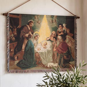 May include: A rectangular woven wall tapestry depicting a nativity scene with figures gathered around a central infant, featuring a fringed edge and suspended from a wooden rod by a thin brown cord, displaying warm golden light and earth tones.