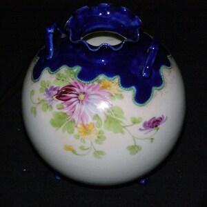 May include: A round, white porcelain vase with a dark blue ruffled rim and two small handles. The vase is decorated with hand-painted flowers in shades of pink, purple, and yellow, with green leaves. The vase has three small feet.