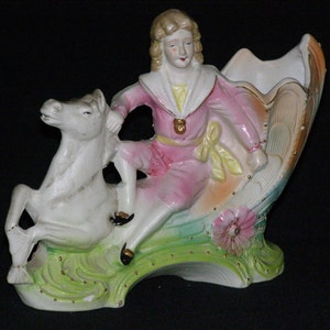 May include: A vintage porcelain figurine of a person riding a white horse. The figure is dressed in a pink outfit with gold accents. The base is decorated with flowers and green grass. The figurine is likely a decorative item.