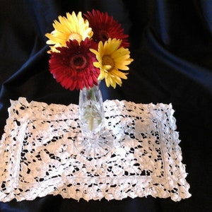 May include: A bouquet of yellow and red gerbera daisies in a clear glass vase on a white lace doily.