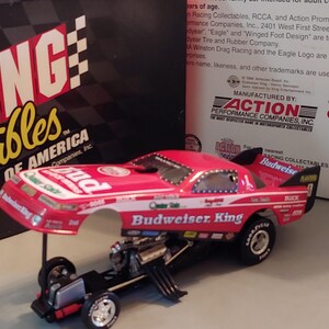 May include: A red and white Budweiser King drag racing collectible car with a black chassis. The car is a Racing Collectibles Club of America product. The box behind the car has a checkered flag design.