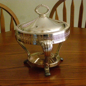 May include: A silver-plated chafing dish with a round lid and three legs. The dish is on a wooden table.