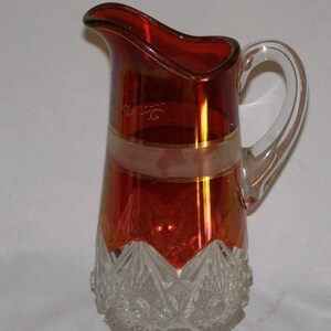 May include: A red glass pitcher with a clear glass handle. The pitcher has a gold band around the middle and a clear glass base with a cut glass design.