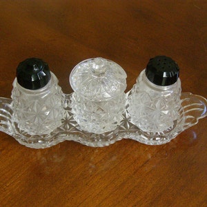May include: A set of three clear glass salt and pepper shakers with black plastic tops, sitting on a clear glass tray with a scalloped edge.