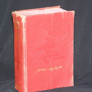 May include: A red hardcover book with gold lettering on the spine. The front cover features a gold embossed design of a quill pen and a wreath. The book is titled "The Complete Works of William Shakespeare".