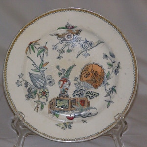 May include: A white ceramic plate with a gold rim and a floral design. The design features a vase with flowers, a fan, and other floral elements in shades of green, blue, red, and yellow.