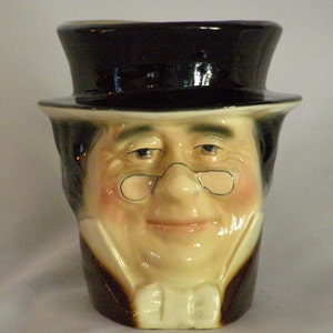 May include: A ceramic mug shaped like a man's head wearing a black top hat and glasses. The mug has a brown and white painted design.